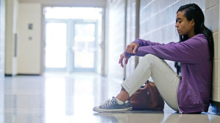 Young girl sitting against white brick wall of a school hallway.